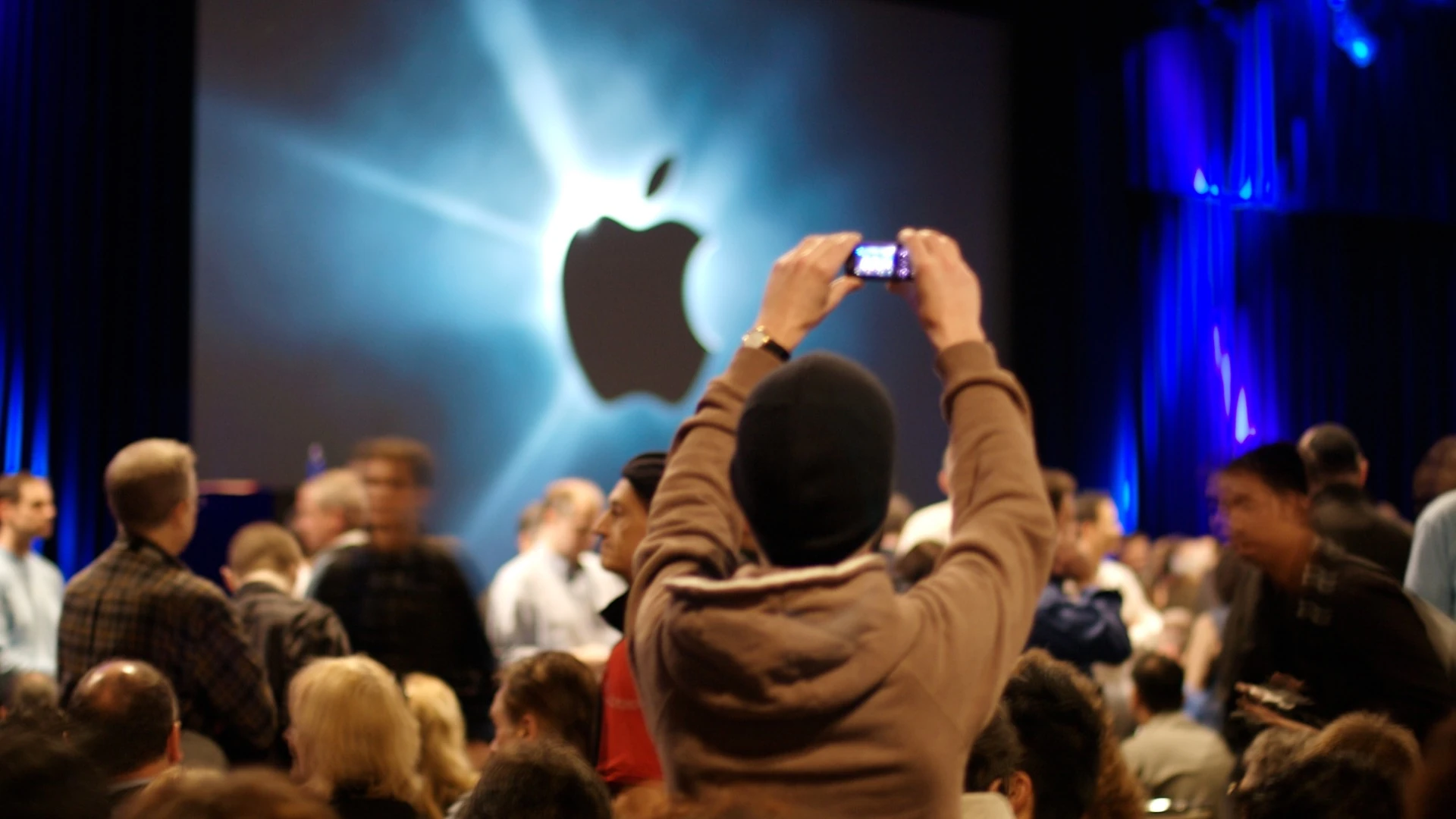 Сцена презентации Apple с крупным светящимся логотипом компании и посетителями в зале
